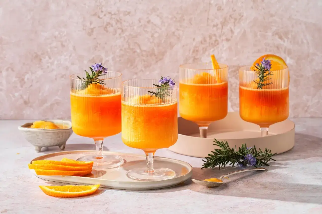 Quattro bicchieri di spritz ghiacciato, un superalcolico rinfrescante, guarniti con rosmarino e fiori viola, su un tavolo con fette d'arancia.