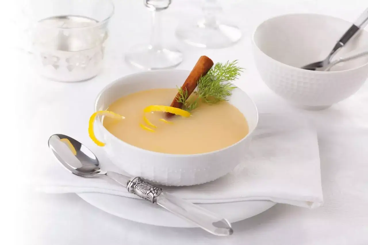 Una ciotola di cremosa salsa al vino bianco, guarnita con scorza di limone, cannella e aneto. Servita con un cucchiaio elegante.