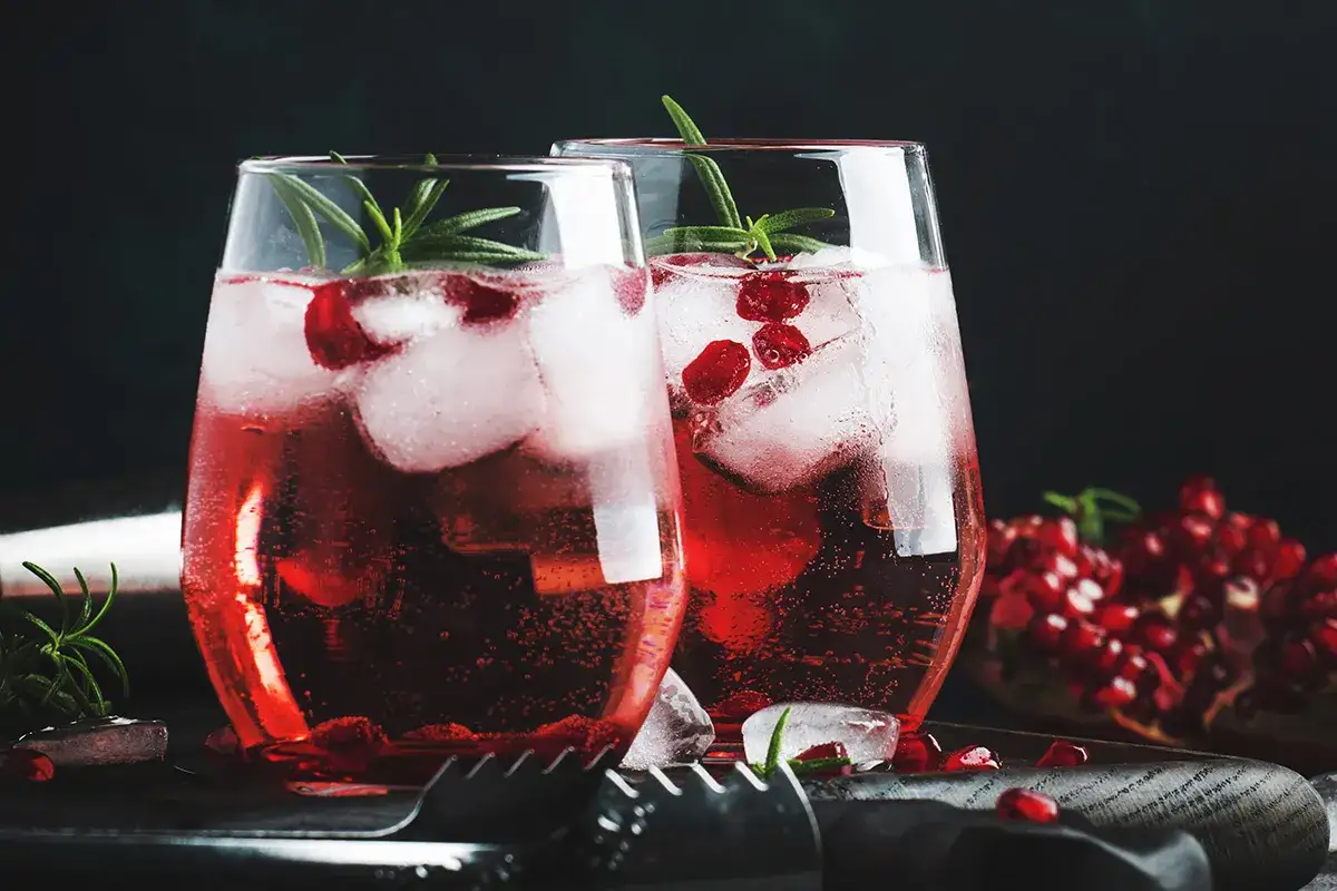 Due bicchieri di spritz al melograno con ghiaccio, chicchi rossi e rosmarino. Sullo sfondo, un melograno aperto.