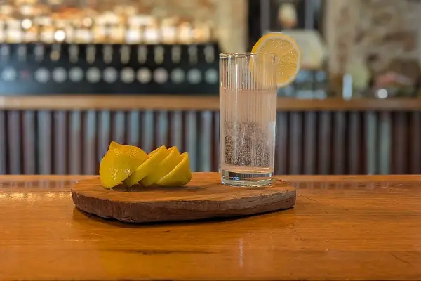 Un bicchiere di acqua frizzante con una fetta di limone, pronto per diventare la base perfetta per un rinfrescante spritz. Accanto, spicchi di limone su un tagliere di legno.